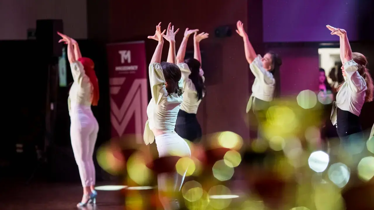 Tancerki w jasnych bluzkach i białych spodniach wykonują zmysłowy taniec. Ich dłonie unoszą się w powietrzu, tworząc dynamiczną choreografię. To piękny pokaz bachata solo.