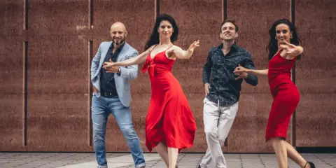 Uśmiechnięci tancerze w czerwonych sukienkach i eleganckich strojach ćwiczą naukę salsy na tle brązowej ściany.