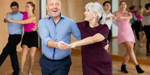 Uśmiechnięta para seniorów uczy się, jak tańczyć na 3, w tle inne pary.