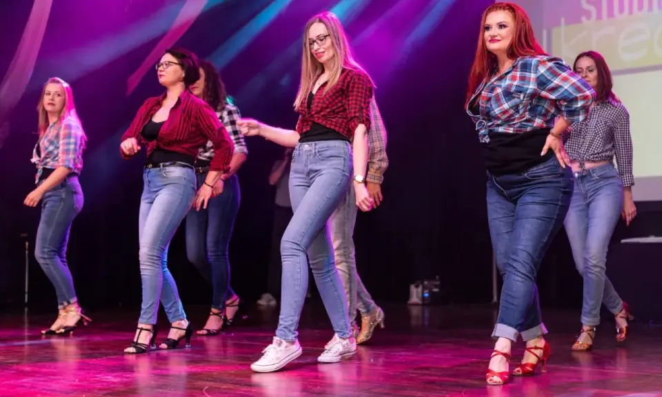 Grupa kobiet w kratę i jeansach tańczy na scenie. Występ latino solo dla początkujących.